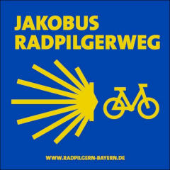 blaues Schild mit gelber Schrift Jakobus Radpilgerweg, mit gelber Jokobsmuschel und gelben Fahrrad
